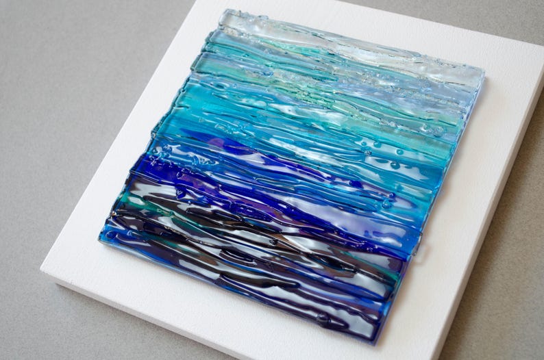 May include: Abstract glass art piece featuring a gradient of blues and greens, resembling ocean waves. The textured glass is mounted on a white square canvas, creating a striking contrast. The artwork evokes a sense of depth and movement.