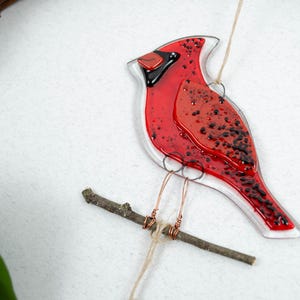 May include: A red and black glass cardinal bird ornament perched on a small wooden branch. The bird has a clear outline and is suspended by a thin string. The cardinal's body is a vibrant red with black speckles.