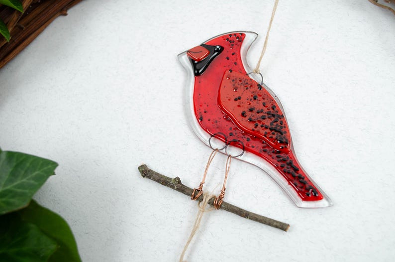 May include: A red and black glass cardinal bird ornament perched on a small wooden branch. The bird has a clear outline and is suspended by a thin string. The cardinal's body is a vibrant red with black speckles.