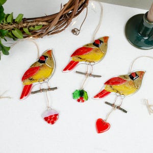 May include: Three decorative cardinal ornaments with red, yellow, and black details, perched on small branches. Each bird hangs from a string, with a red heart, holly, and a red and white ornament suspended below. A candle holder and wreath are in the background.