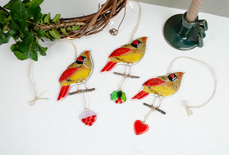 May include: Three decorative cardinal ornaments with red, yellow, and black details, perched on small branches. Each bird hangs from a string, with a red heart, holly, and a red and white ornament suspended below. A candle holder and wreath are in the background.