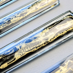 May include: Four clear glass tiles with black and gold marbling inside. The tiles are arranged in a row, with the top tile slightly offset from the others.