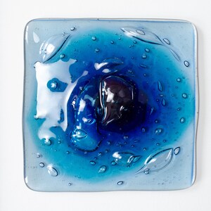 May include: A square fused glass art piece featuring a gradient of blue tones, ranging from light cyan to deep navy. The surface is textured with numerous small air bubbles and organic, fluid shapes, all encased within a simple black frame.