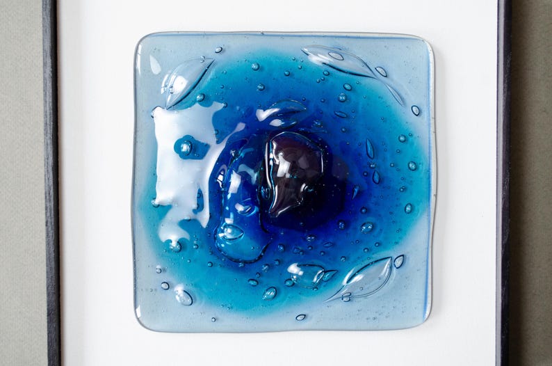 May include: A square fused glass art piece featuring a gradient of blue tones, ranging from light cyan to deep navy. The surface is textured with numerous small air bubbles and organic, fluid shapes, all encased within a simple black frame.