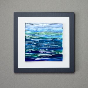 May include: Abstract art piece featuring layered glass in shades of blue and green, resembling a seascape. The artwork is mounted on a white background within a dark blue frame, creating a modern aesthetic. The piece is square in shape.