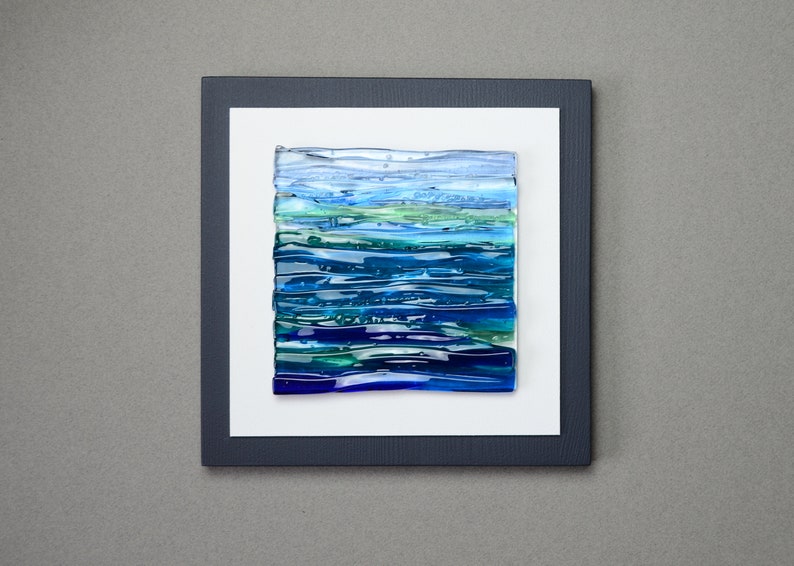 May include: Abstract art piece featuring layered glass in shades of blue and green, resembling a seascape. The artwork is mounted on a white background within a dark blue frame, creating a modern aesthetic. The piece is square in shape.