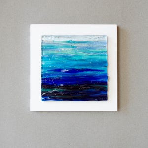 May include: Abstract ocean art piece. The artwork features a square of layered glass in shades of blue, teal, and white, mounted on a white square. The colors blend to depict a seascape, with dark blues at the bottom and lighter hues at the top.