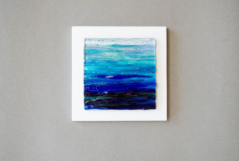 May include: Abstract ocean art piece. The artwork features a square of layered glass in shades of blue, teal, and white, mounted on a white square. The colors blend to depict a seascape, with dark blues at the bottom and lighter hues at the top.