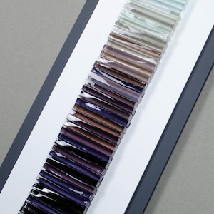 May include: A sample card of glass rods in various shades of purple, brown, and gray. The rods are arranged in a vertical line, with the darkest shades at the bottom and the lightest shades at the top.