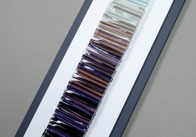 May include: A sample card of glass rods in various shades of purple, brown, and gray. The rods are arranged in a vertical line, with the darkest shades at the bottom and the lightest shades at the top.