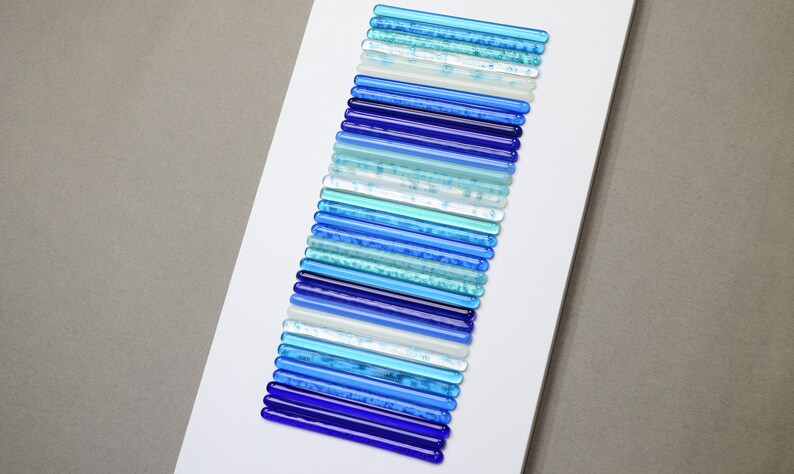 Pu&ograve; includere: Un'opera d'arte da parete realizzata con bacchette di vetro in varie tonalit&agrave; di blu, dal blu chiaro al blu scuro, disposte in uno schema verticale.