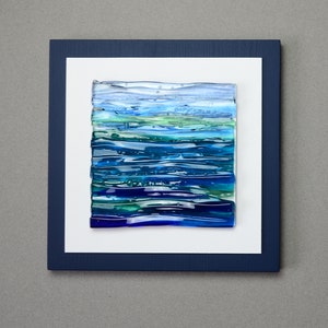 May include: Abstract glass art piece with layered blue, green, and clear glass, mounted on a white mat and framed in a navy blue frame. The artwork evokes a sense of water or landscape.