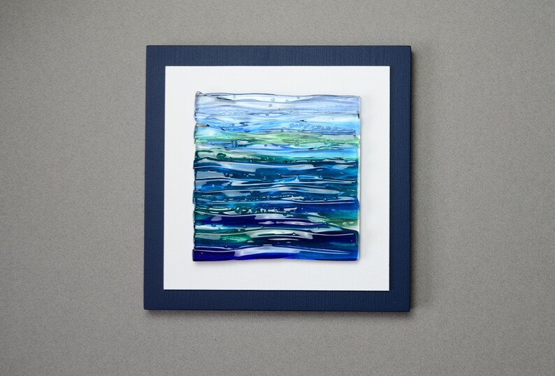 May include: Abstract glass art piece with layered blue, green, and clear glass, mounted on a white mat and framed in a navy blue frame. The artwork evokes a sense of water or landscape.