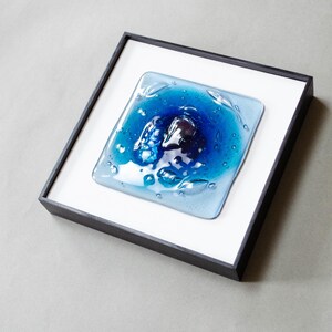 May include: A square fused glass art piece featuring a deep blue center that transitions to lighter blue edges, set within a black frame against a white mat. The glass surface has a textured, rippled appearance with visible air bubbles throughout.