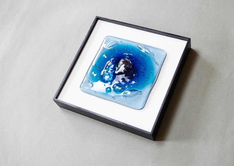 May include: A square fused glass art piece featuring a deep blue center that transitions to lighter blue edges, set within a black frame against a white mat. The glass surface has a textured, rippled appearance with visible air bubbles throughout.