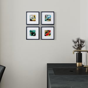 May include: A set of four square wall art pieces in black frames with white matting, each featuring abstract colorful glass designs. The artwork is displayed on a plain wall above a dark kitchen counter with a black and gold faucet and a small plant.
