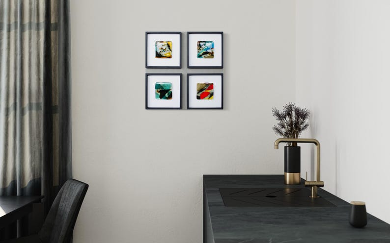 May include: A set of four square wall art pieces in black frames with white matting, each featuring abstract colorful glass designs. The artwork is displayed on a plain wall above a dark kitchen counter with a black and gold faucet and a small plant.