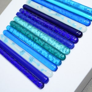 May include: A wall art piece made of glass rods in various shades of blue, from light blue to dark blue, arranged in a gradient pattern. The glass rods have a textured surface and are arranged in a vertical line.