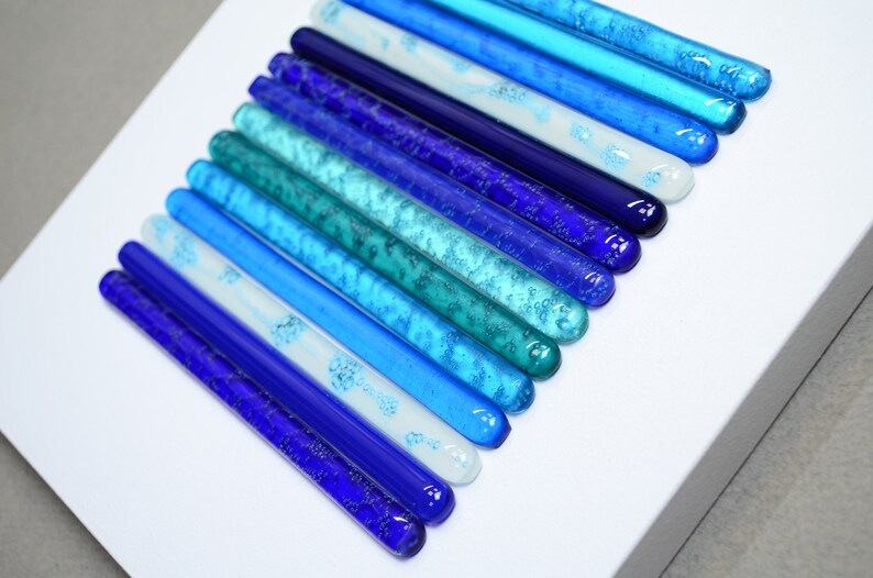 May include: A wall art piece made of glass rods in various shades of blue, from light blue to dark blue, arranged in a gradient pattern. The glass rods have a textured surface and are arranged in a vertical line.