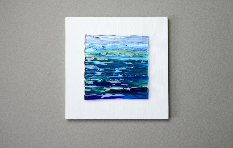 May include: Abstract art piece featuring a rectangular composition of layered glass in shades of blue and green, resembling a seascape. The artwork is mounted on a white, textured square canvas, creating a striking contrast against the neutral gray background.