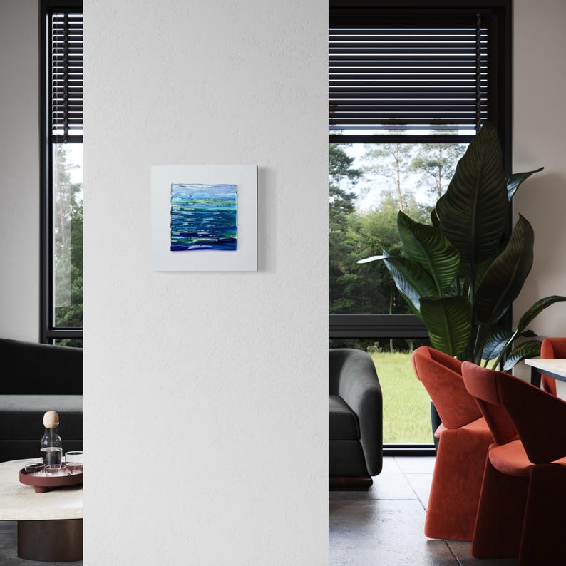 May include: Square abstract painting with a white border. The artwork features horizontal brushstrokes in shades of blue and green, creating a textured, water-like effect. The painting is mounted on a white wall in a modern interior.