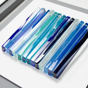 Pu&ograve; includere: Un'opera d'arte da parete incorniciata che presenta un design a strati di strisce di vetro blu, turchese e bianco. Le strisce di vetro sono disposte in un motivo verticale, creando un aspetto moderno e astratto.