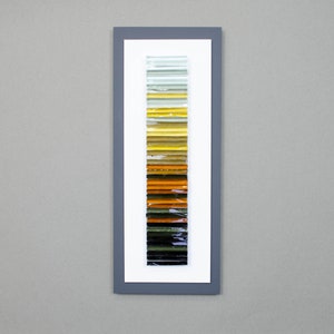 May include: A framed artwork featuring a vertical arrangement of colorful glass tiles. The tiles transition from light green to yellow, orange, brown, and black.