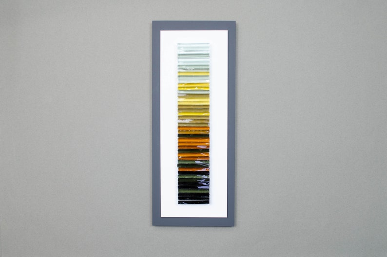 May include: A framed artwork featuring a vertical arrangement of colorful glass tiles. The tiles transition from light green to yellow, orange, brown, and black.