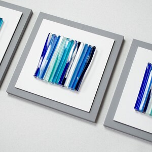 Pu&ograve; includere: Tre pezzi di arte da parete incorniciati con un design astratto moderno. Ogni pezzo presenta una disposizione verticale di bacchette di vetro blu, turchese e bianche.