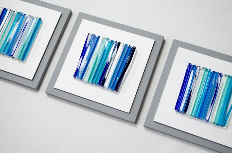 Pu&ograve; includere: Tre pezzi di arte da parete incorniciati con un design astratto moderno. Ogni pezzo presenta una disposizione verticale di bacchette di vetro blu, turchese e bianche.