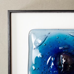 May include: A square fused glass art piece featuring a gradient of deep cobalt to light turquoise blue. The surface is textured with numerous small air bubbles and raised, leaf-like glass elements, all encased within a thin black frame.
