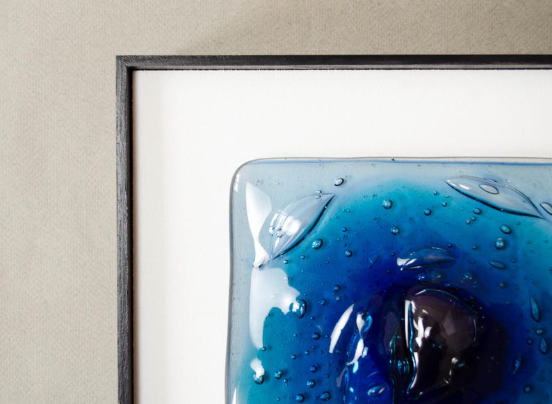 May include: A square fused glass art piece featuring a gradient of deep cobalt to light turquoise blue. The surface is textured with numerous small air bubbles and raised, leaf-like glass elements, all encased within a thin black frame.