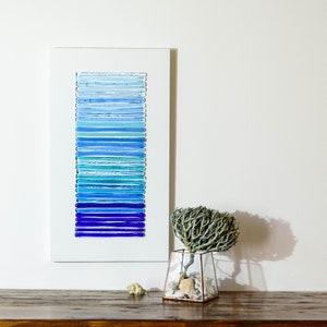 May include: A framed wall art piece featuring a gradient of blue glass tiles. The art is displayed on a wooden shelf with a small vase of dried branches and seashells.