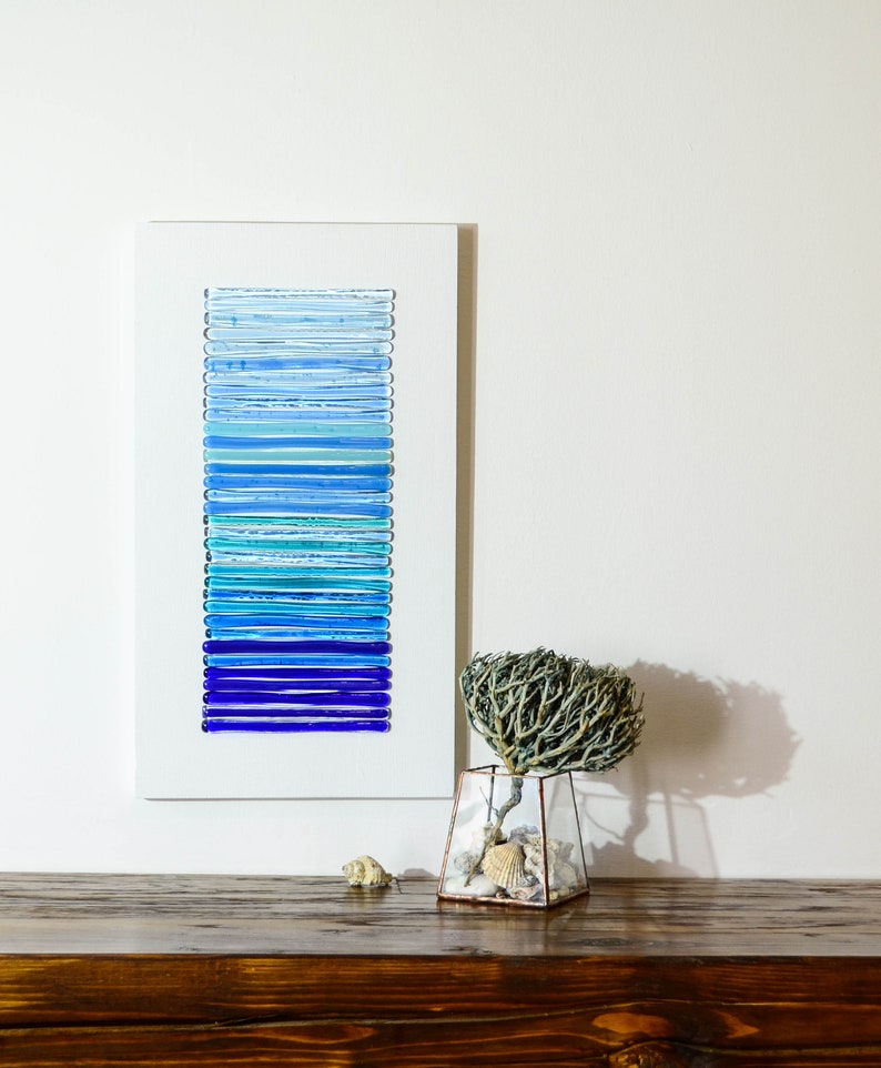 May include: A framed wall art piece featuring a gradient of blue glass tiles. The art is displayed on a wooden shelf with a small vase of dried branches and seashells.