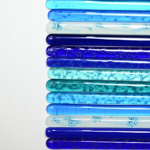 May include: A collection of glass rods in various shades of blue, teal, and white, arranged in a gradient pattern. The rods have a textured surface with small air bubbles trapped inside.