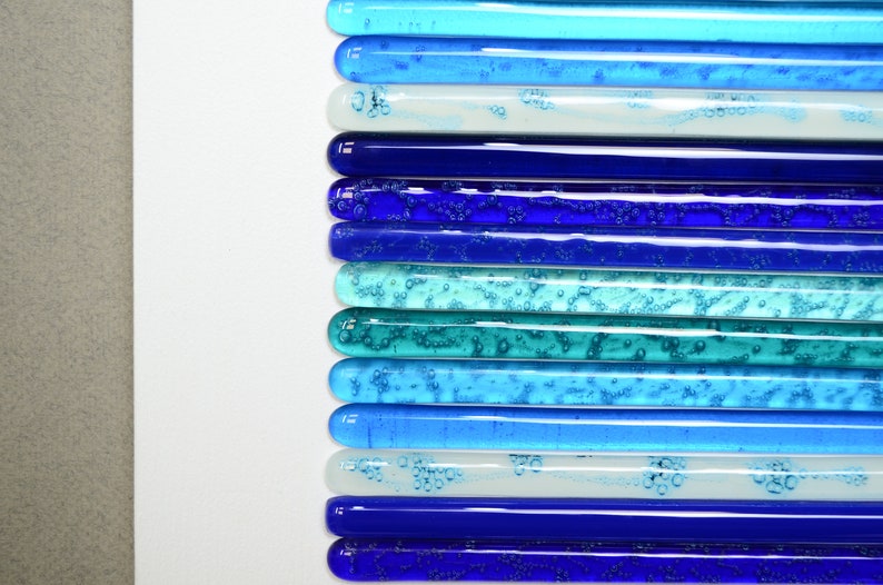 May include: A collection of glass rods in various shades of blue, teal, and white, arranged in a gradient pattern. The rods have a textured surface with small air bubbles trapped inside.