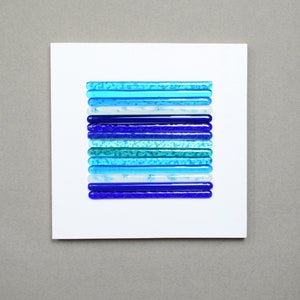 May include: A white card with a blue and white glass mosaic art piece. The mosaic is made of small, rectangular pieces of glass in shades of blue, turquoise, and white. The pieces are arranged in horizontal rows, creating a gradient effect from dark blue to light blue.