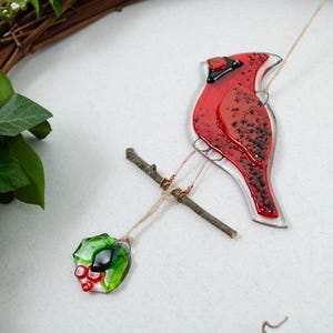 May include: A red glass cardinal bird ornament with black accents, perched on a small twig. The bird is suspended by a string, with a green and red glass holly berry ornament hanging below. A natural wreath is in the background.