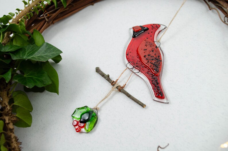 May include: A red glass cardinal bird ornament with black accents, perched on a small twig. The bird is suspended by a string, with a green and red glass holly berry ornament hanging below. A natural wreath is in the background.