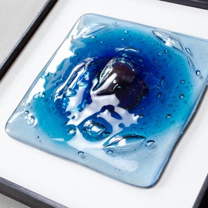 May include: A square fused glass art piece featuring a gradient of blue tones, ranging from deep navy to light sky blue. The surface has a glossy, textured finish with visible air bubbles trapped within the glass, all set inside a black frame.