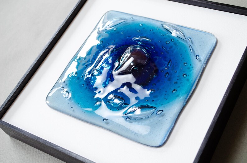 May include: A square fused glass art piece featuring a gradient of blue tones, ranging from deep navy to light sky blue. The surface has a glossy, textured finish with visible air bubbles trapped within the glass, all set inside a black frame.