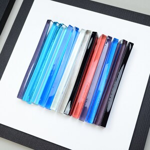 Pu&ograve; includere: Un'opera d'arte da parete incorniciata che presenta una collezione di bacchette di vetro colorate disposte in uno schema verticale. Le bacchette sono in tonalit&agrave; di blu, bianco, nero, rosso, viola e grigio.