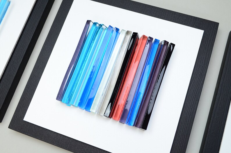 Pu&ograve; includere: Un'opera d'arte da parete incorniciata che presenta una collezione di bacchette di vetro colorate disposte in uno schema verticale. Le bacchette sono in tonalit&agrave; di blu, bianco, nero, rosso, viola e grigio.