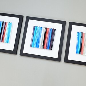 Pu&ograve; includere: Tre pezzi di arte da parete quadrati incorniciati con sfondo bianco e cornici nere. Ogni pezzo presenta una disposizione verticale di bacchette di vetro colorate in tonalit&agrave; di blu, rosso, bianco e grigio.