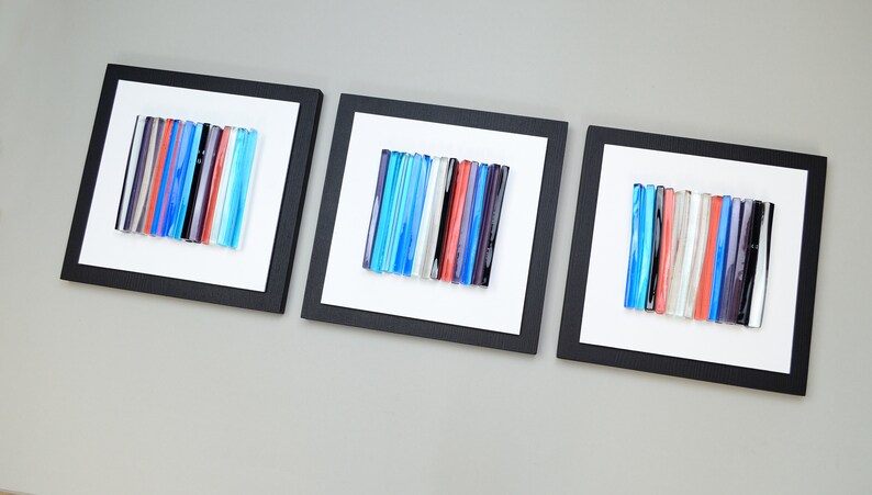Pu&ograve; includere: Tre pezzi di arte da parete quadrati incorniciati con sfondo bianco e cornici nere. Ogni pezzo presenta una disposizione verticale di bacchette di vetro colorate in tonalit&agrave; di blu, rosso, bianco e grigio.