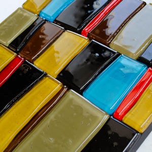 May include: A mosaic tile art piece with a geometric pattern of rectangular tiles in various colours. The tiles include shades of yellow, brown, black, red, blue, and olive green, arranged diagonally.