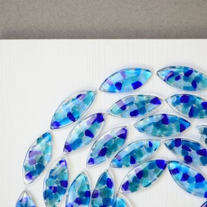 May include: A circular arrangement of blue and turquoise glass tiles, each with a unique pattern of smaller glass pieces, on a white background.