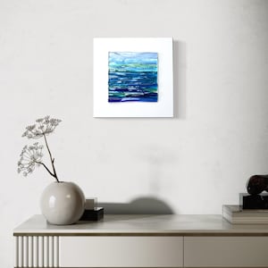 May include: Abstract art piece featuring a square composition of layered blue and green hues, reminiscent of water. The artwork is set within a white frame, creating a modern aesthetic. The piece is displayed on a white wall.