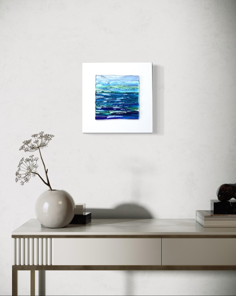 May include: Abstract art piece featuring a square composition of layered blue and green hues, reminiscent of water. The artwork is set within a white frame, creating a modern aesthetic. The piece is displayed on a white wall.