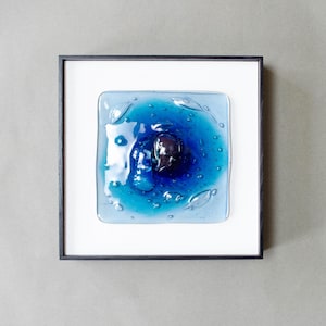 May include: A square wall art piece featuring a fused glass panel with shades of blue, ranging from light to deep navy, centered with a dark, rounded glass element. The glass has a rippled, liquid-like texture and is set in a thin black frame.